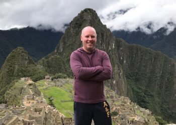 Don Trynor standing on a lookout during his Machu Picchu science travel experience, with the ancient citadel behind him and surrounded by steep mountain cliffs and drifting clouds.