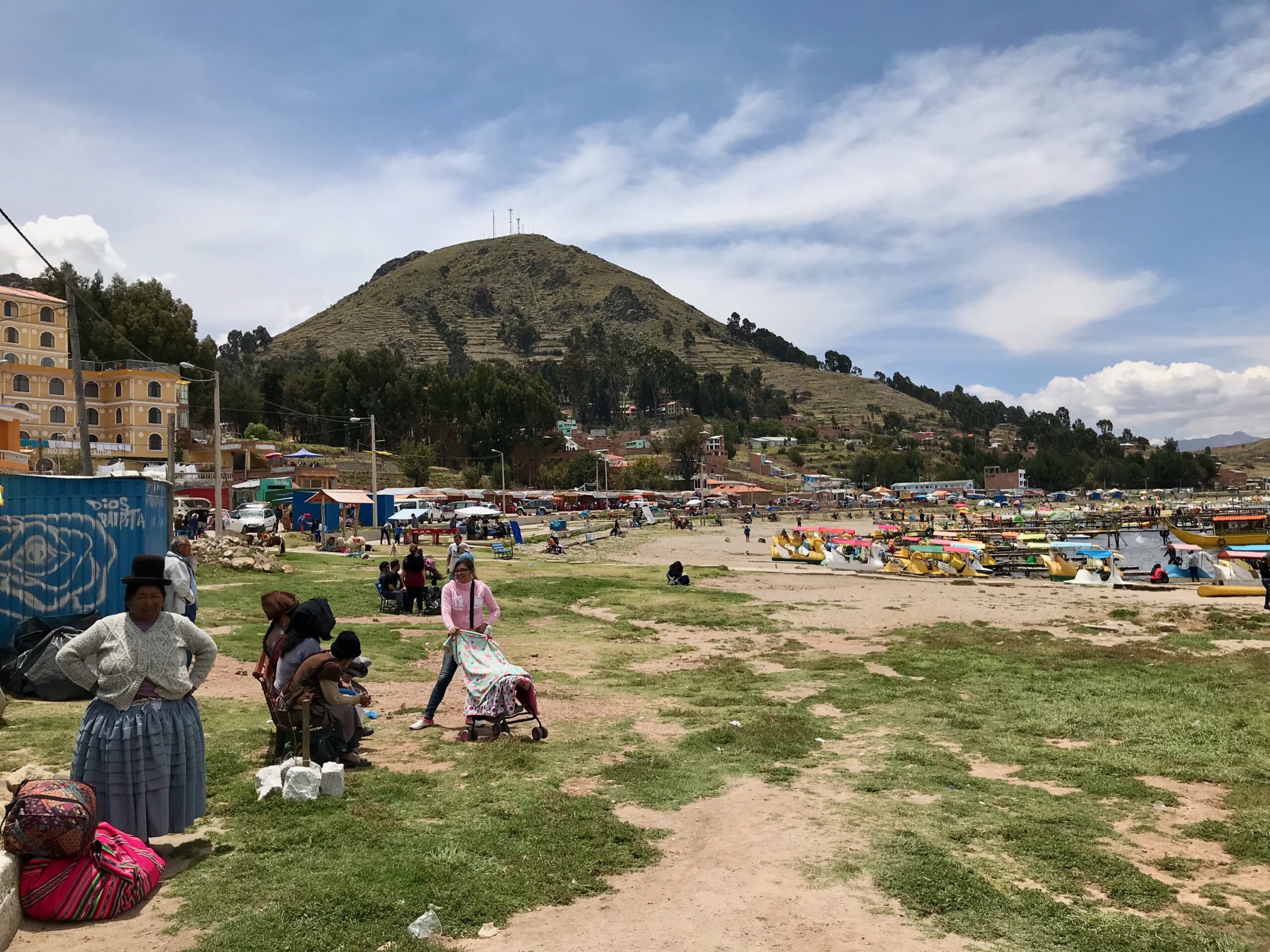 Lakeside area of Copacabana on Lake Titicaca with paddle boats and families in traditional clothing picnicking by the shore.