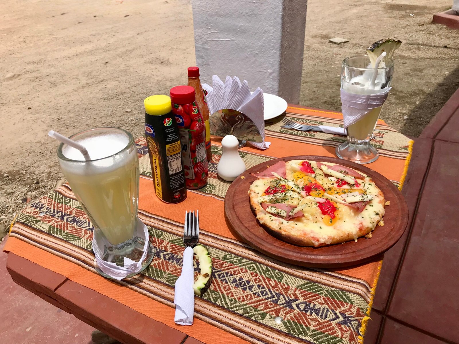 Table set with a pizza topped with vegetables and ham alongside glasses of fresh pineapple juice, photographed in Copacabana, Bolivia, near Lake Titicaca.