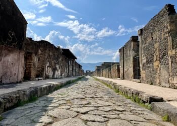 Photo of Pompeii showing a cobblestone road stretching into the distance, flanked by ancient buildings, with mountains visible on the horizon.