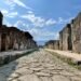 Photo of Pompeii showing a cobblestone road stretching into the distance, flanked by ancient buildings, with mountains visible on the horizon.