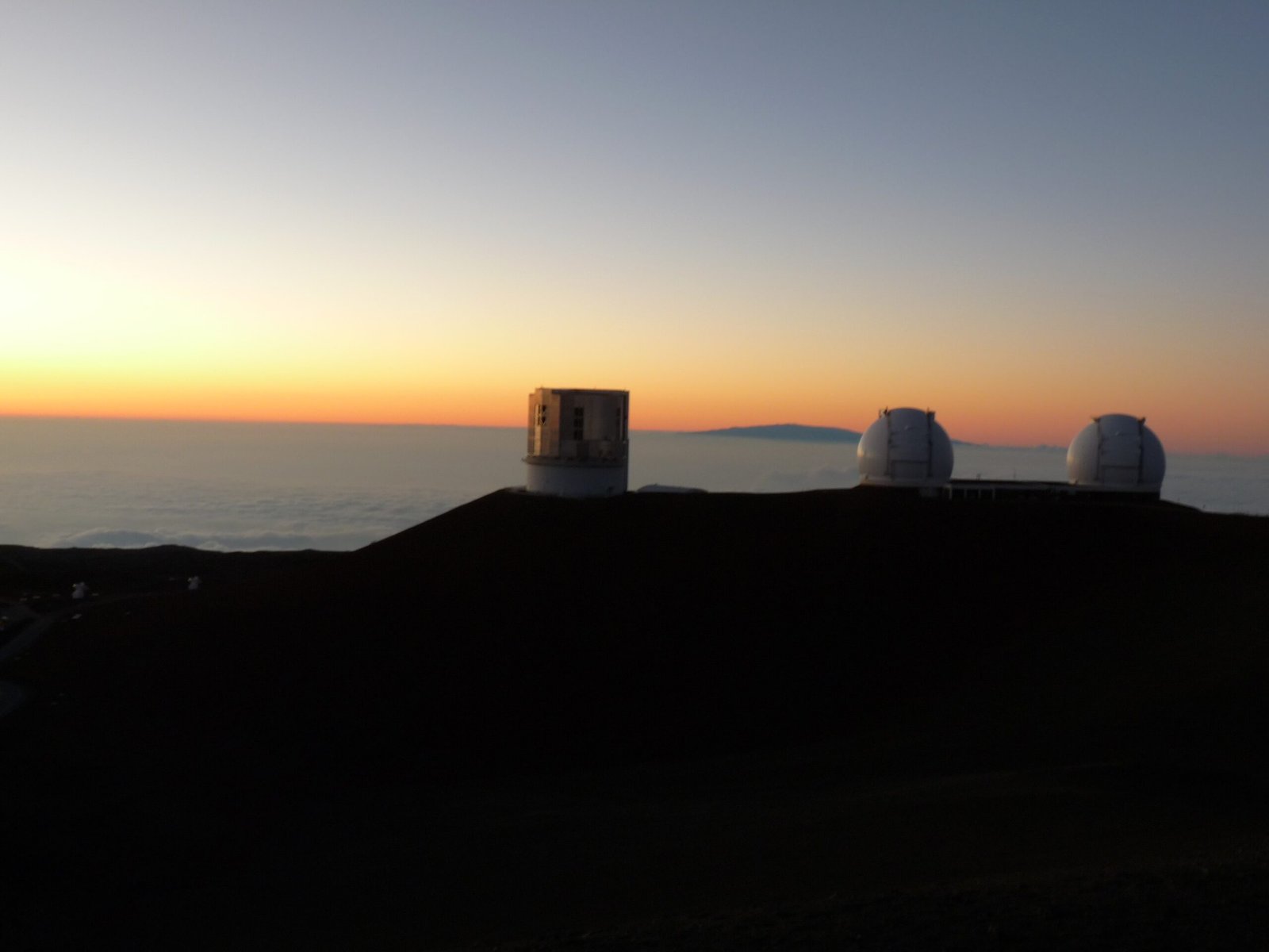 Mauna Kea summit at twilight with several observatories visible and a sea of clouds below the volcanic peak.