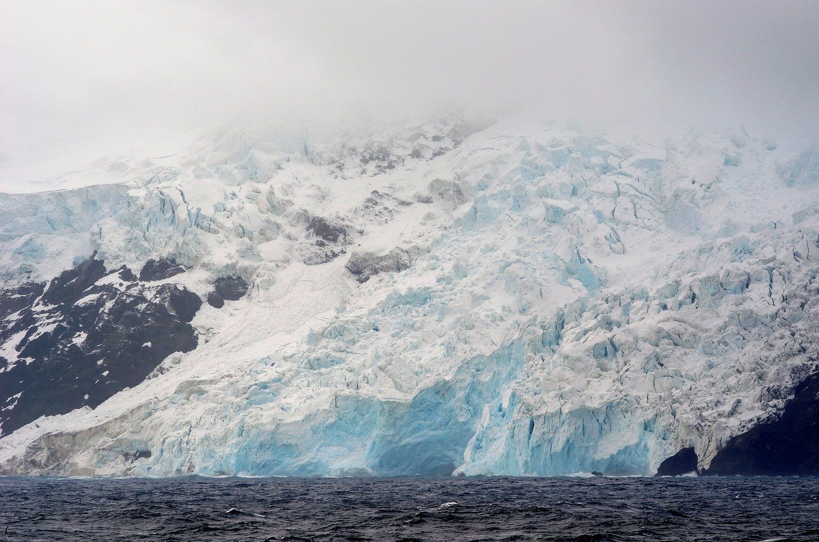A glacier on the west coast of Bouvet Island, featuring icy cliffs and rugged terrain under a cloudy sky. Photo by Fguerraz, via Wikimedia Commons.