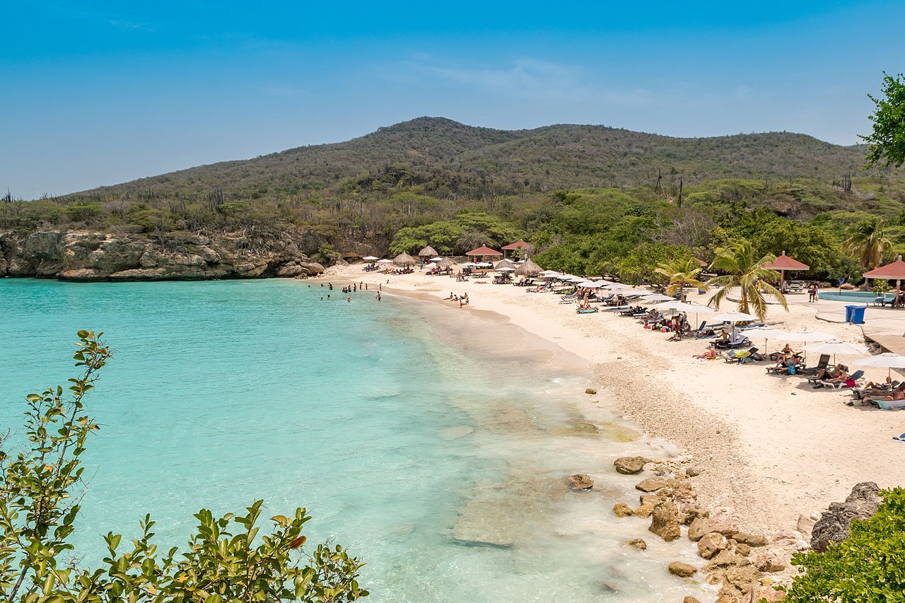 A scenic beach in Curacao with turquoise blue water, sunbathers on the sandy shore, and clear skies. This location is also a favorite ham radio vacation destination for ham radio operators seeking tropical paradise. Photo by dronepicr, licensed under CC BY 2.0.