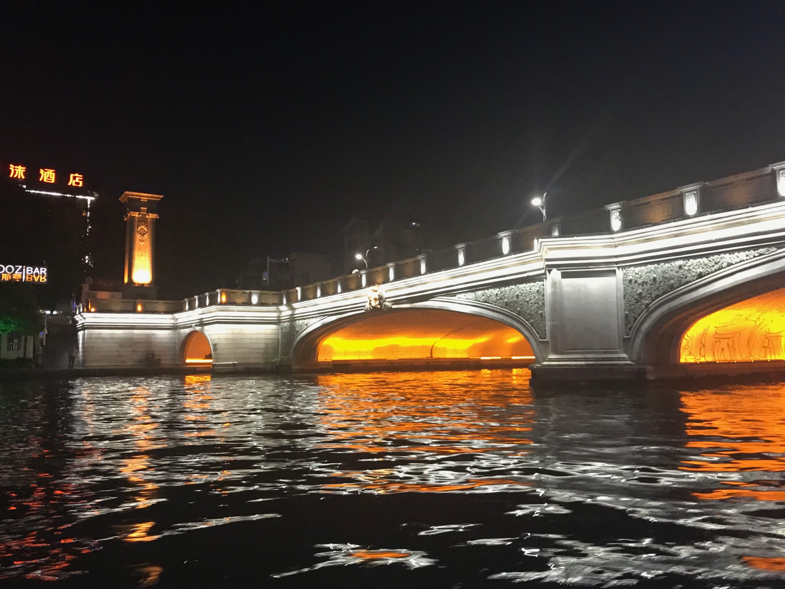 Colorfully lit bridge spanning the Li River in Guilin during a night cruise, glowing in white and orange lights that reflect on the water.