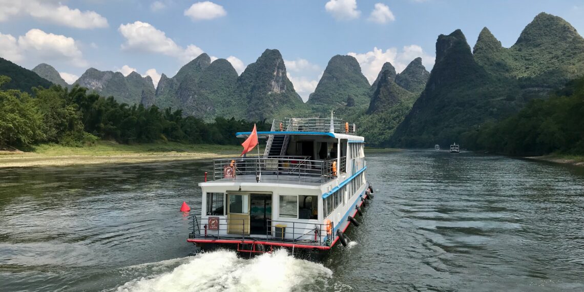 A river cruise boat on the Li River in Guilin, China, with towering limestone peaks in the background against a bright sky.