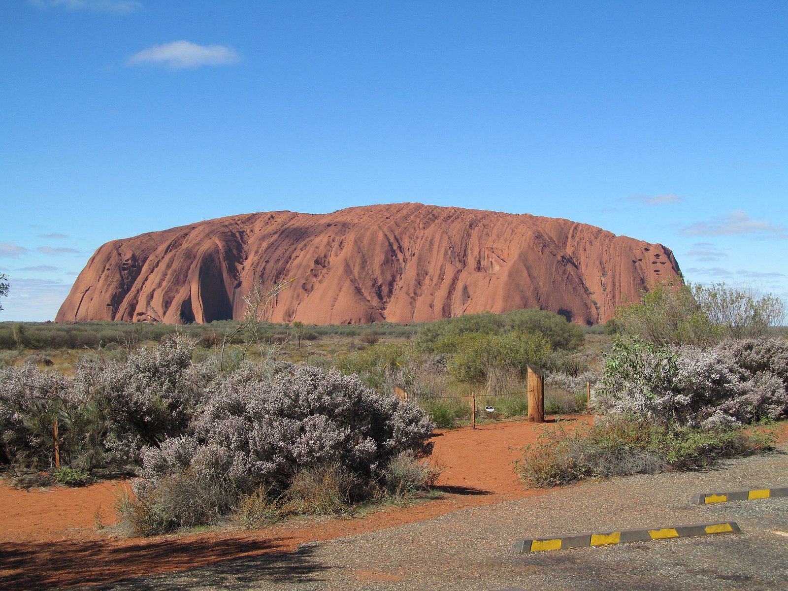 Uluru in the Australian Outback, a massive red sandstone monolith surrounded by sparse vegetation under a clear blue sky. Photo by Broome2, via Wikimedia Commons.