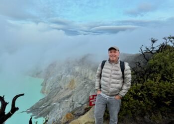 Don Trynor, author at Curious Don, stands on the rim of the Kawah Ijen volcano crater in Indonesia, with a turquoise crater lake visible below.