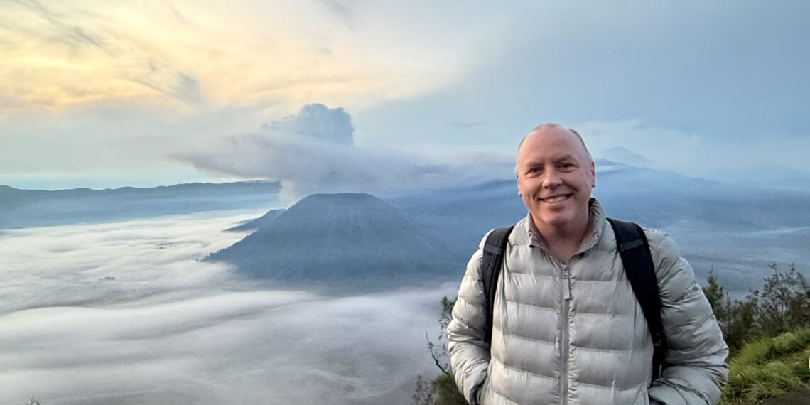 Don Trynor standing at Bukit Cinta at dawn, with the smoking Mount Bromo volcano rising above a mist-filled Tengger Caldera in the background.