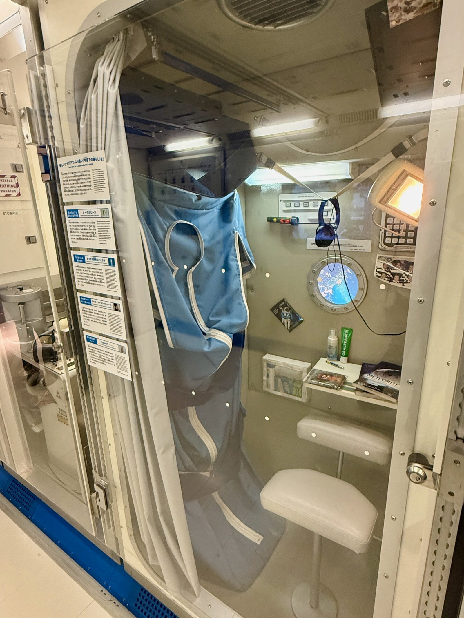 Interior of a replica ISS crew sleeping quarters at Miraikan, showing a sleeping bag, shelf with books and toiletries, and a small chair.