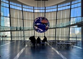Silhouetted visitors sitting at a table in front of the illuminated Geo-Cosmos globe in Miraikan’s main hall.