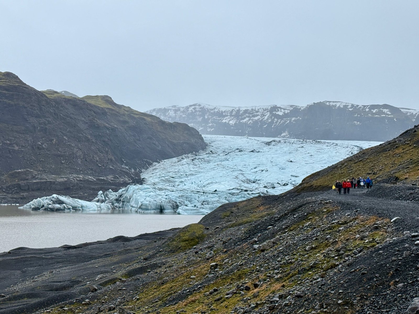 A distant view of Sólheimajökull glacier in Iceland, descending between snow-capped mountains toward a lagoon, with black volcanic ash and rocky terrain in the foreground.