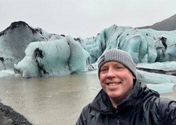 A photo of Don Trynor in front of Solheimajokull glacier, showing blue-tinted ice covered in black volcanic ash.