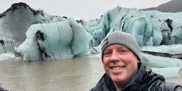 A photo of Don Trynor in front of Solheimajokull glacier, showing blue-tinted ice covered in black volcanic ash.