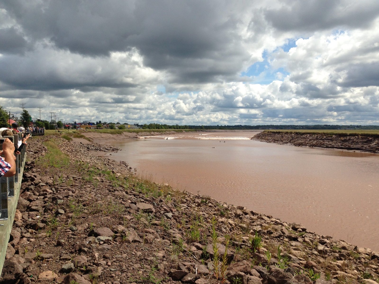 The Moncton tidal bore rounds a bend in the chocolate-brown Petitcodiac River, with surfers riding the wave near Chateau Moncton on a sunny August day.