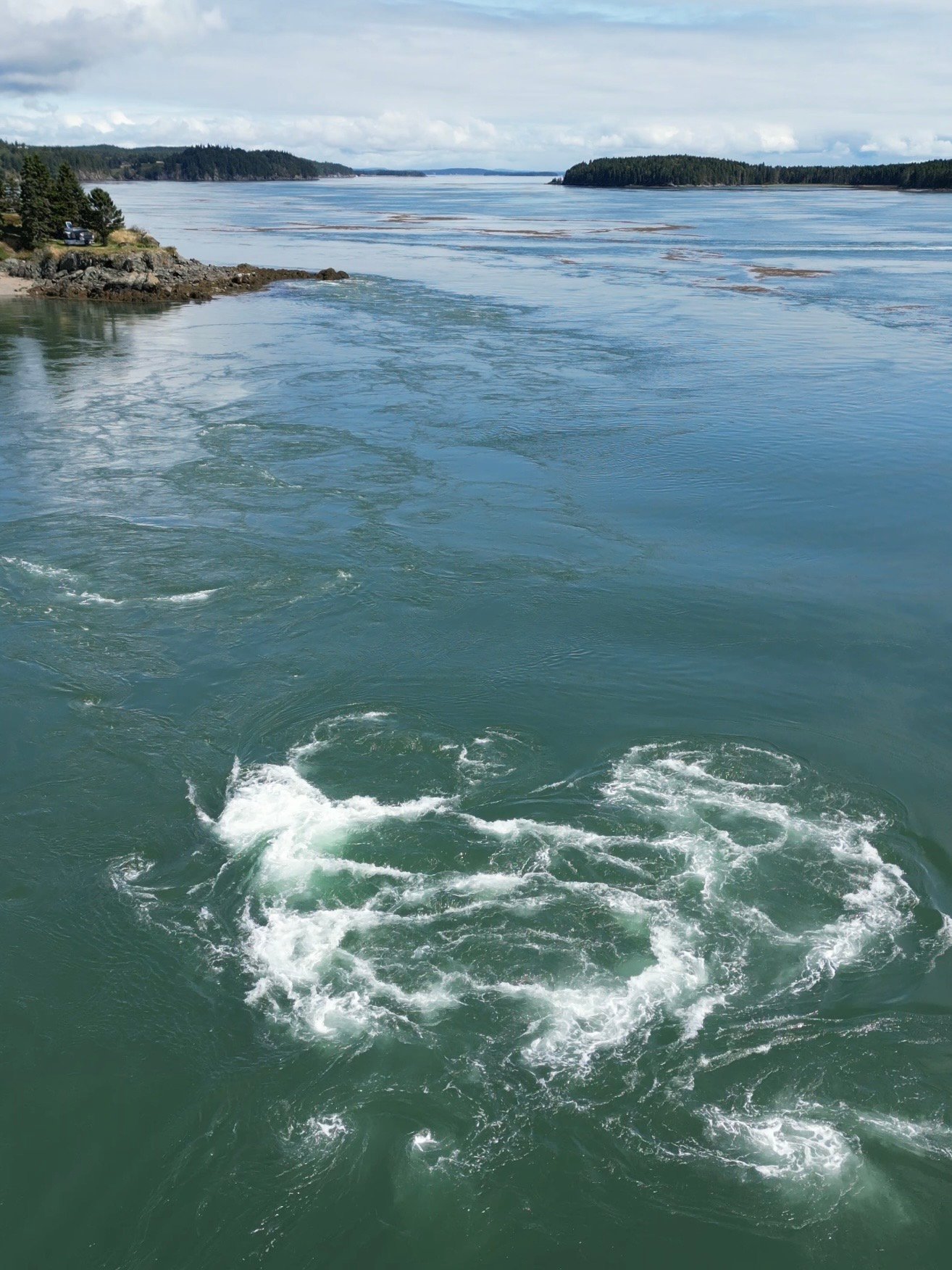 A view of the Western Passage near Deer Island Point Light Beacon, showing swirling upwelling water and foam patterns formed by the Old Sow Whirlpool’s strong tidal currents against a backdrop of forested islands.