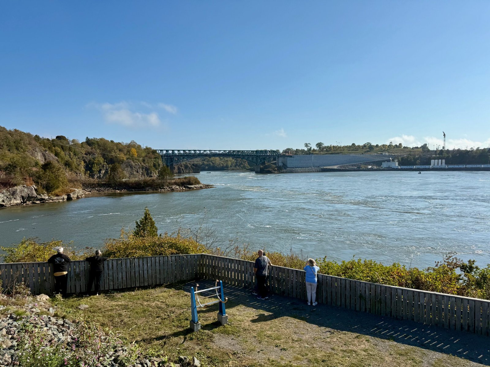 A group of visitors standing along the wooden fence at Fallsview Park in Saint John, New Brunswick, overlooking the Reversing Falls as the Bay of Fundy tide floods upriver beneath the Reversing Falls Bridge.