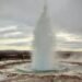 The Strokkur Geyser erupting in Iceland, with boiling water shooting into the air and steam rising around it.