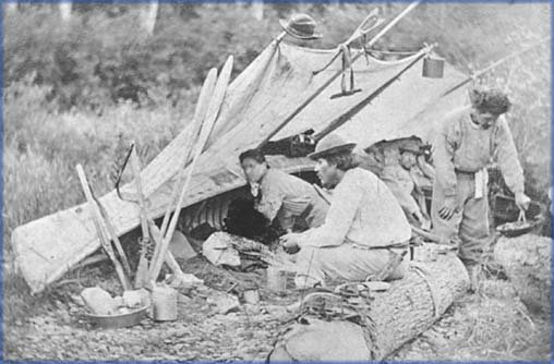 Historic 1865 photo of Wolastoqiyik (Maliseet) people at a Blue Mountain Camp on the Tobique River, showing canoes, shelters, rifles, paddles, and cooking utensils.