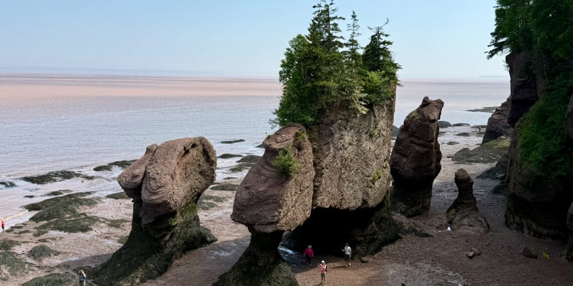 Visitors walking beneath the rock formations at Hopewell Rocks during the Hopewell Rocks tides at low tide, with the Bay of Fundy in the background.