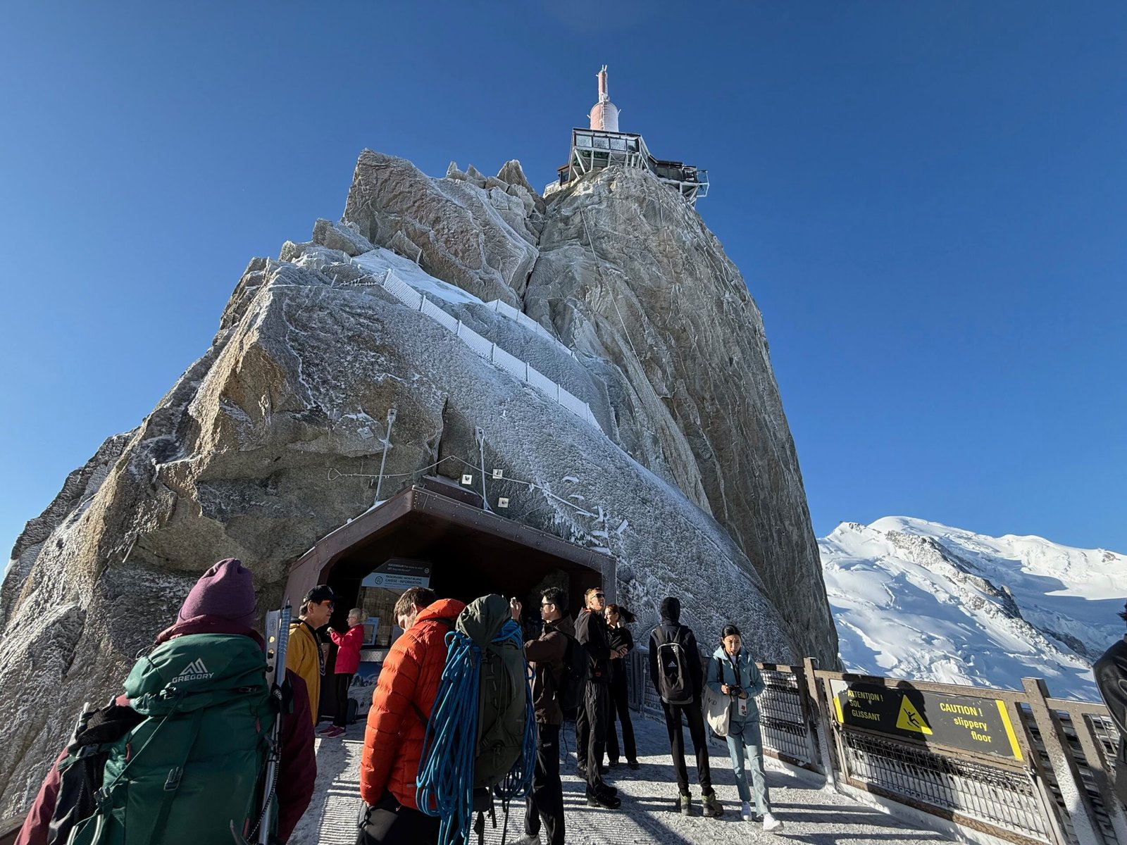 View from the Aiguille du Midi summit station showing the frost-covered broadcast antenna on the granite peak, with Mont Blanc rising snow-covered in the background and visitors admiring the scene.