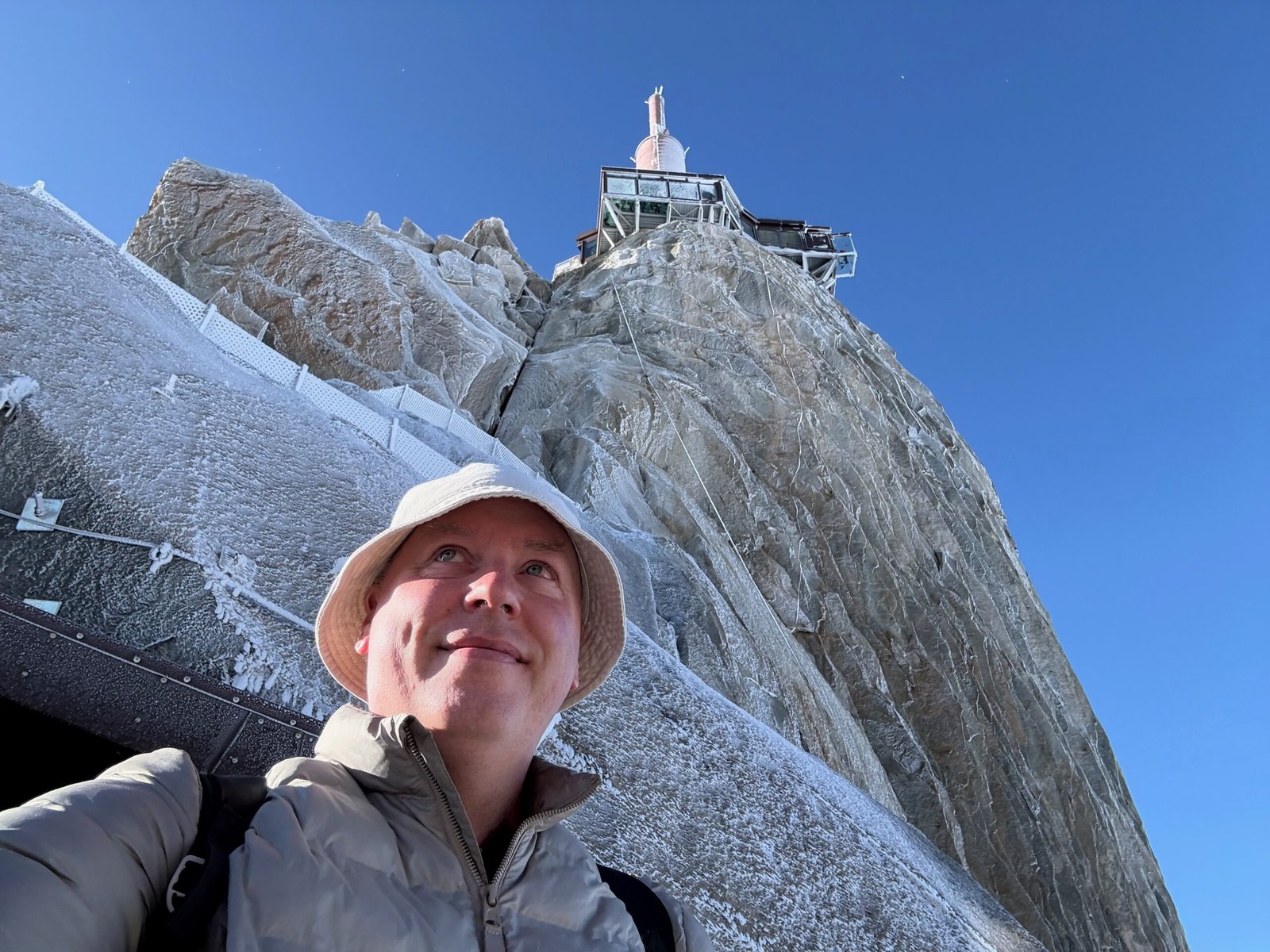 Don Trynor, author of Curious Don, at the Aiguille du Midi summit station with the upper level and broadcast antenna visible against a clear blue sky.