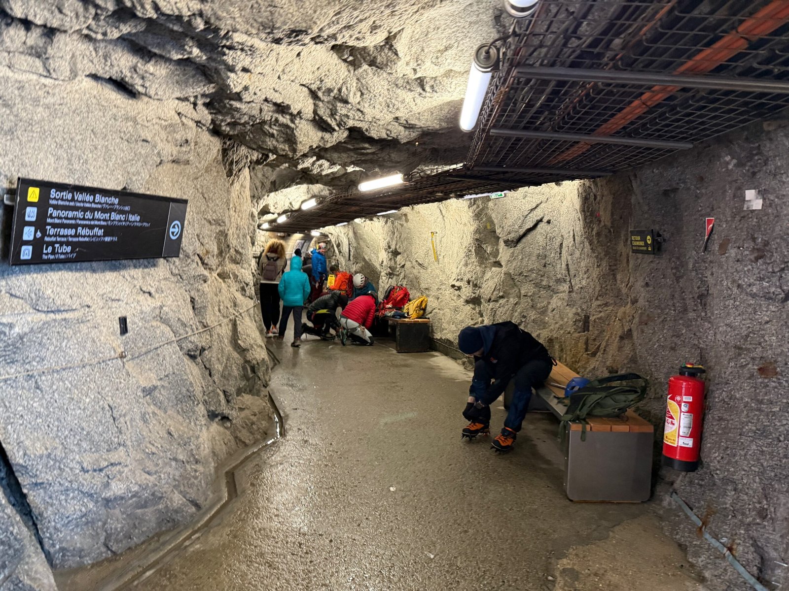 Inside a granite tunnel at the Aiguille du Midi with visitors preparing to hike, showing bare granite walls and ceiling carved into the mountain.