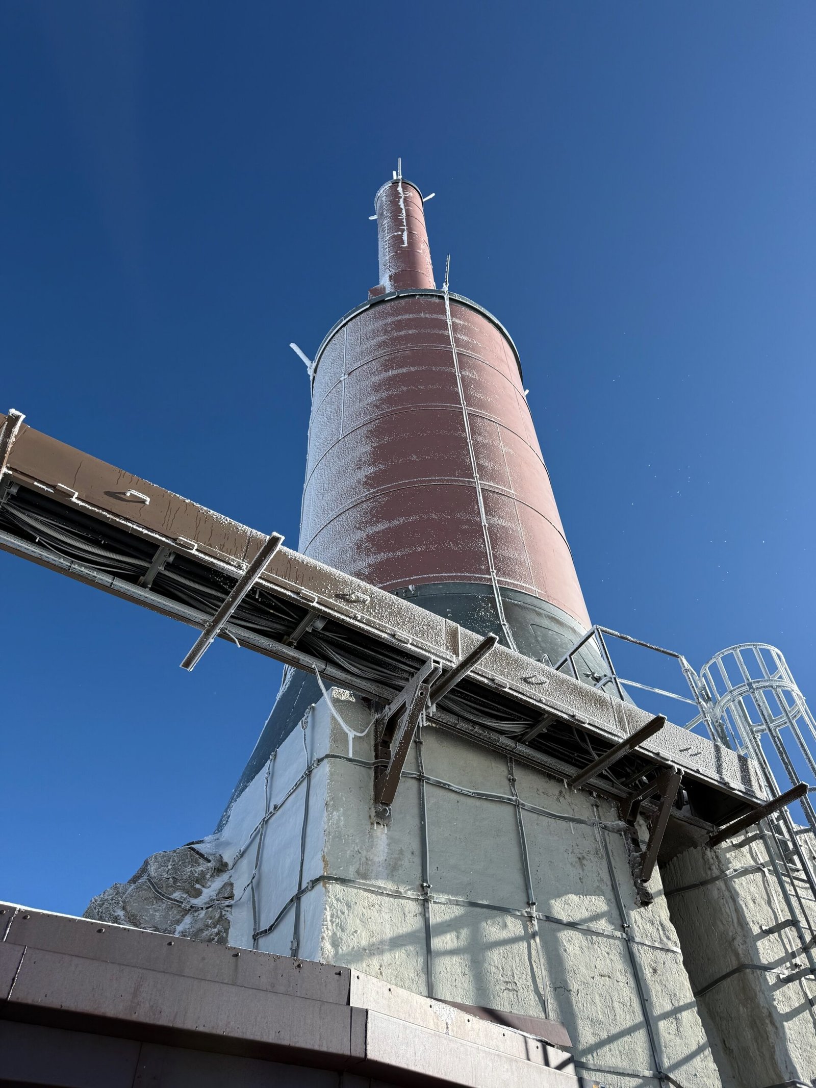 Broadcast antenna at the summit of the Aiguille du Midi, covered in frost against a clear blue sky, used for television and FM radio transmission.