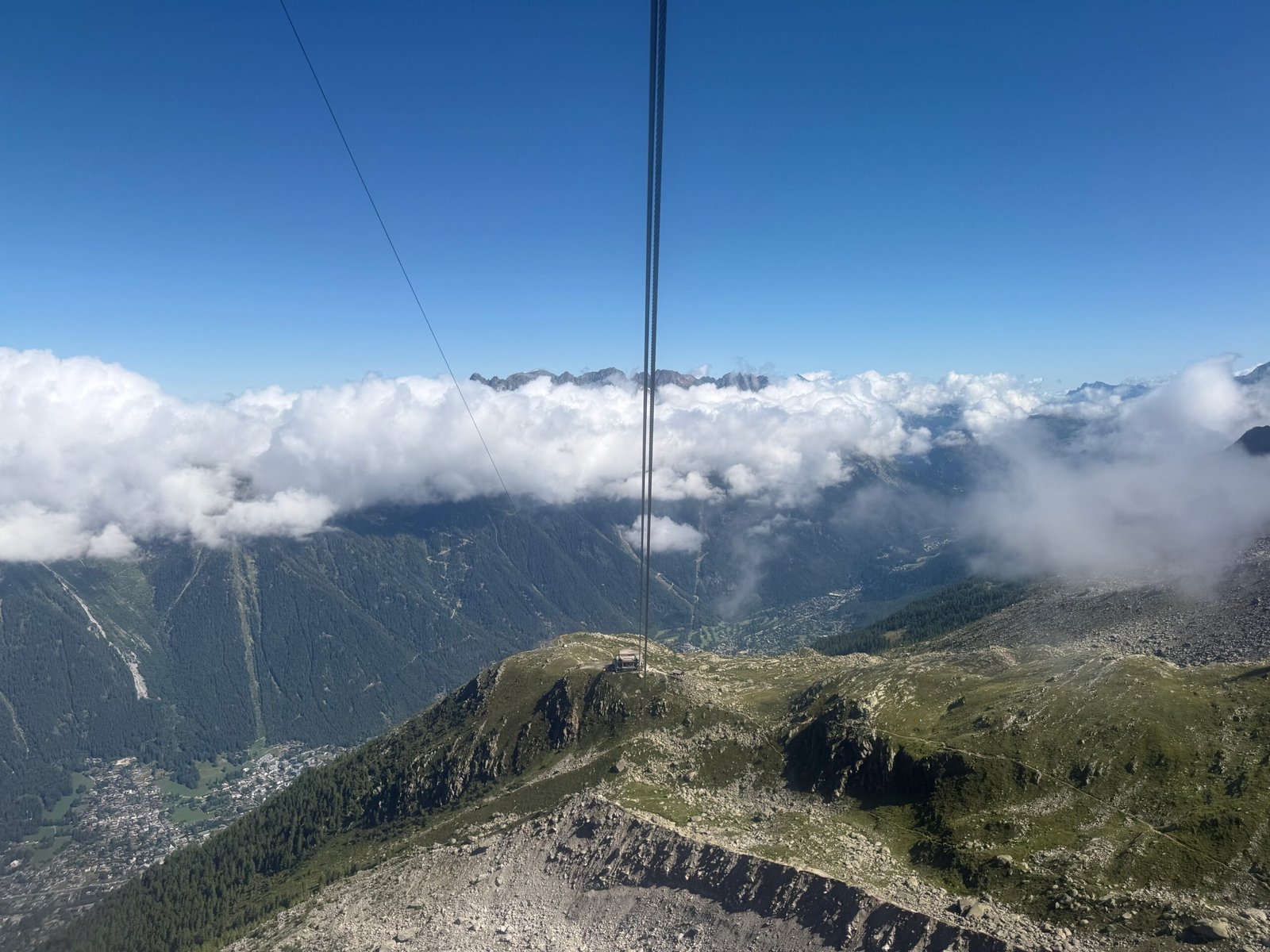 View from the Aiguille du Midi cable car descending toward the Plan de l’Aiguille mid-station, with Chamonix in the valley below and clouds drifting over the Alps.
