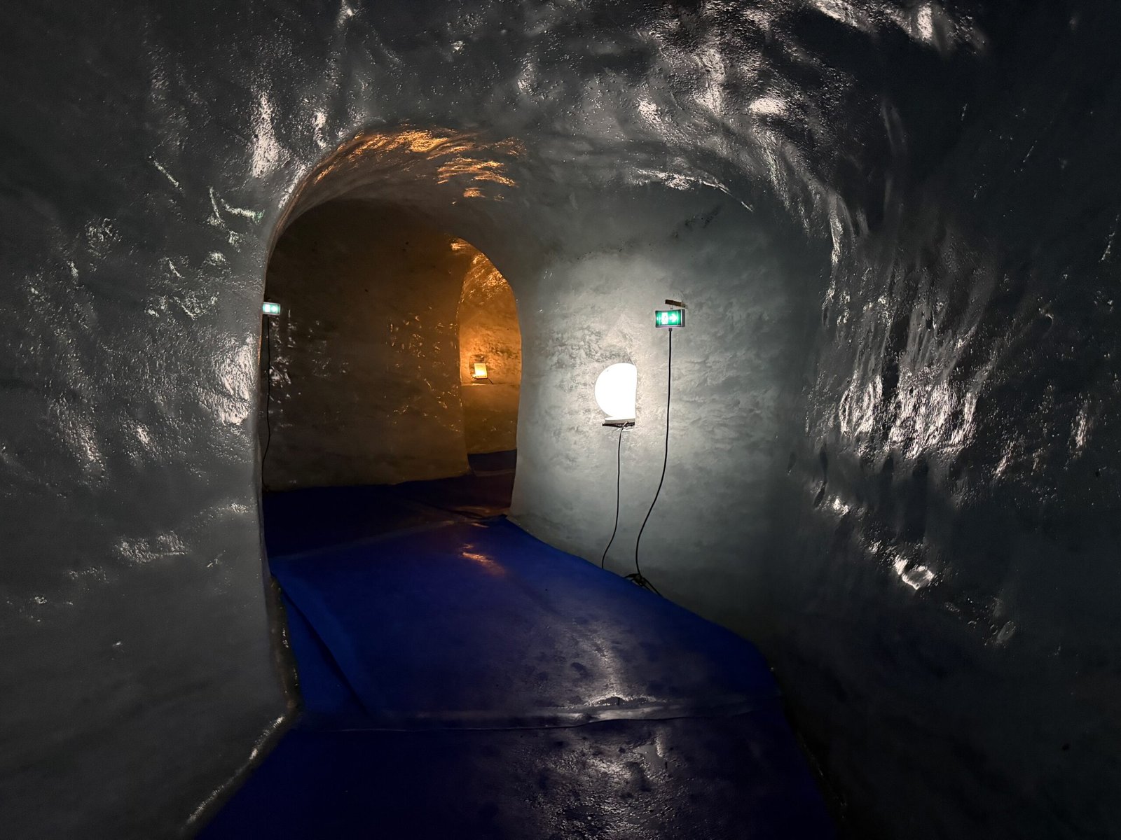 Dimly lit ice tunnel inside the Grotte de Glace, with lamps illuminating glistening ice walls and a glowing orange chamber visible through an arched corridor.