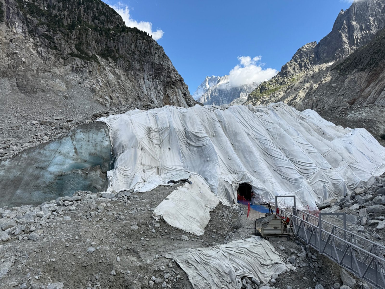 Grotte de Glace entrance on the Mer de Glace glacier covered in white thermal tarps, with rocky terrain and mountains in the background.
