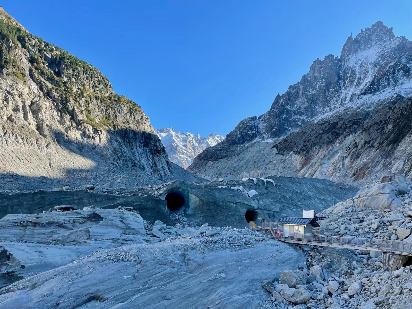 View of Mer de Glace glacier in October 2021 showing multiple old Grotte de Glace entrances stacked along the ice face, with rocky valley and alpine peaks in background.