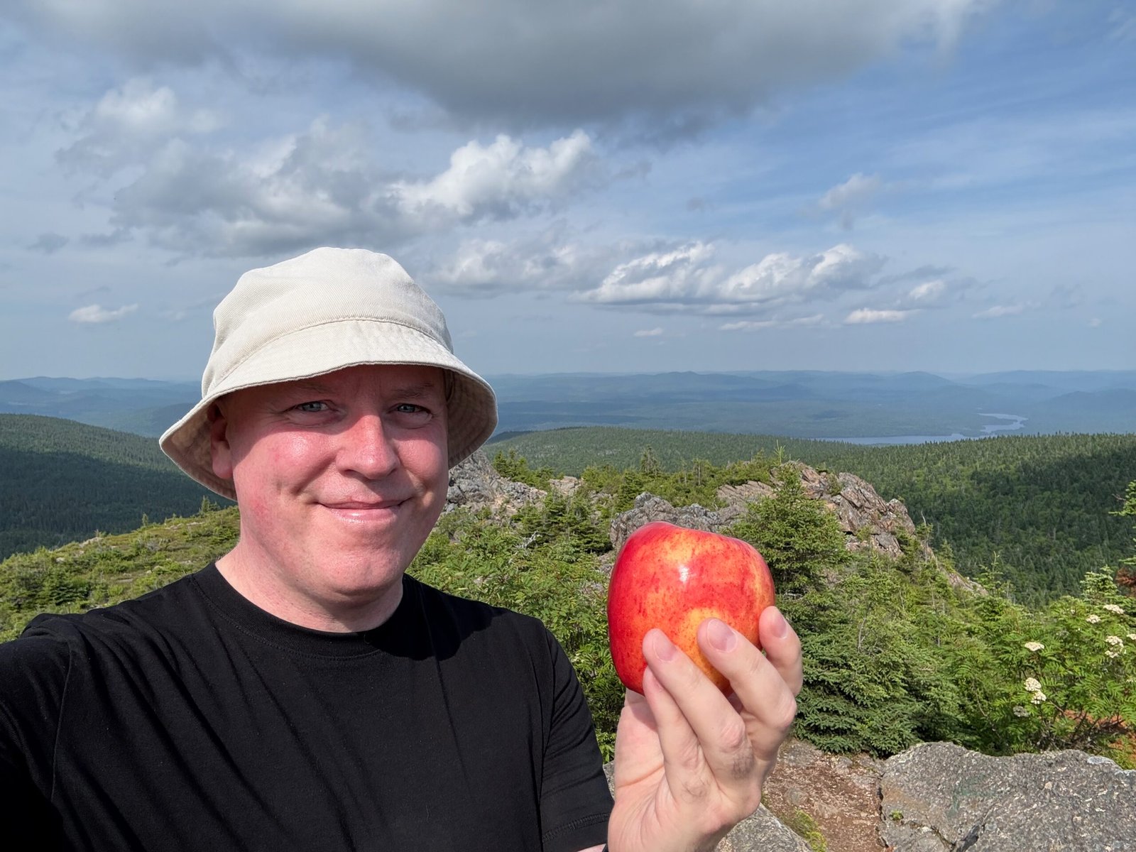 Hiker enjoying a snack with an apple at the summit of Mount Carleton, overlooking Nictau Lake and the Appalachian Mountains in New Brunswick.