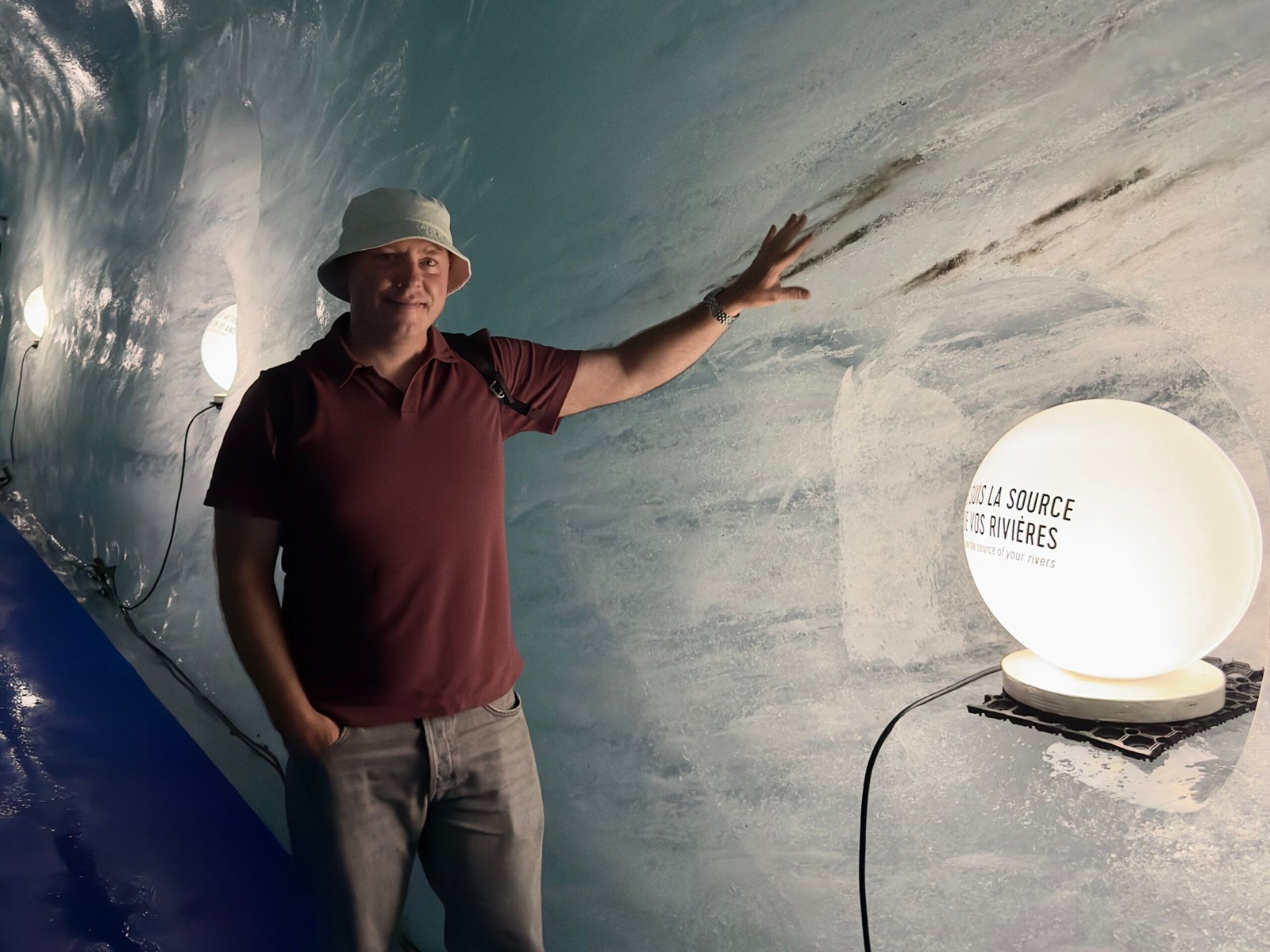 Don Trynor standing inside the Grotte de Glace, the Mer de Glace ice cave, with his hand on the icy wall beside an illuminated educational lamp.