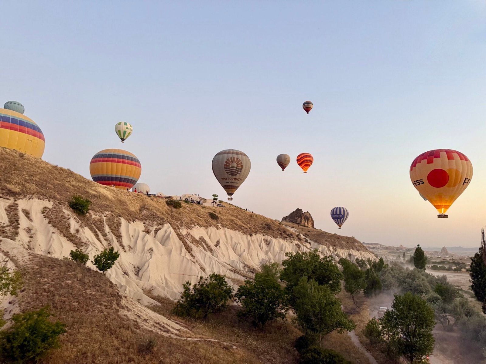 Colourful Cappadocia hot air balloons rising at dawn above rocky cliffs and green valleys, seen from a low cruising balloon in Türkiye.