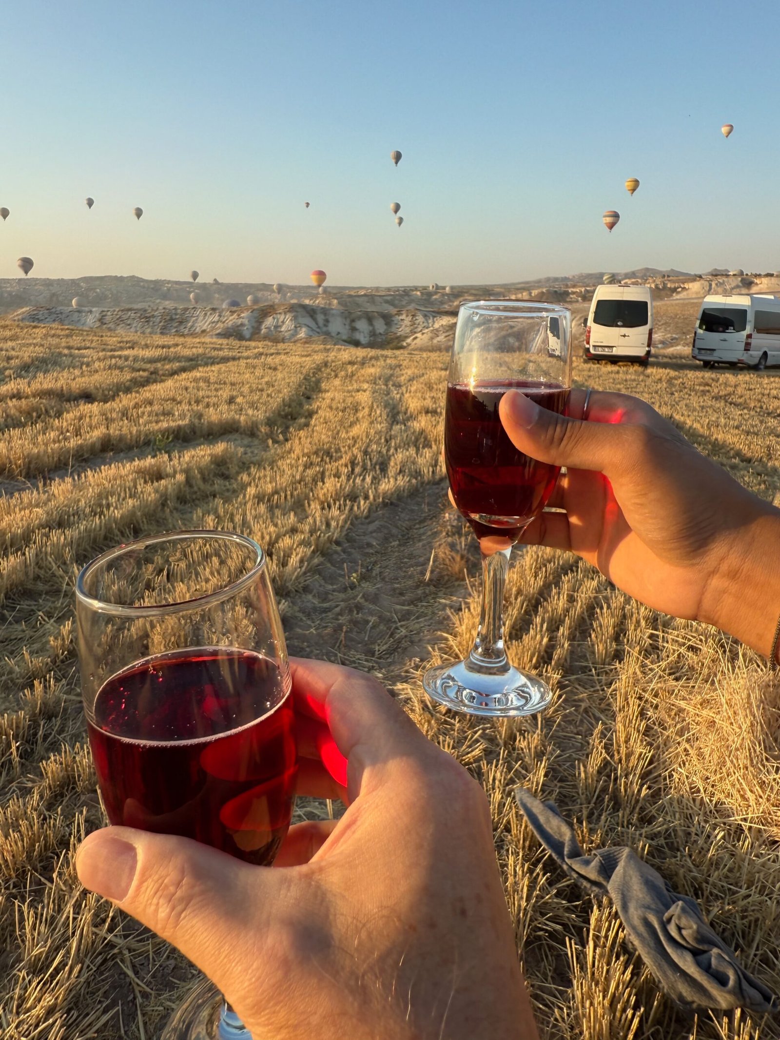 Two glasses of champagne and cranberry juice held aloft for a toast after a Cappadocia hot air balloon landing in Türkiye, with balloons still visible in the morning sky.