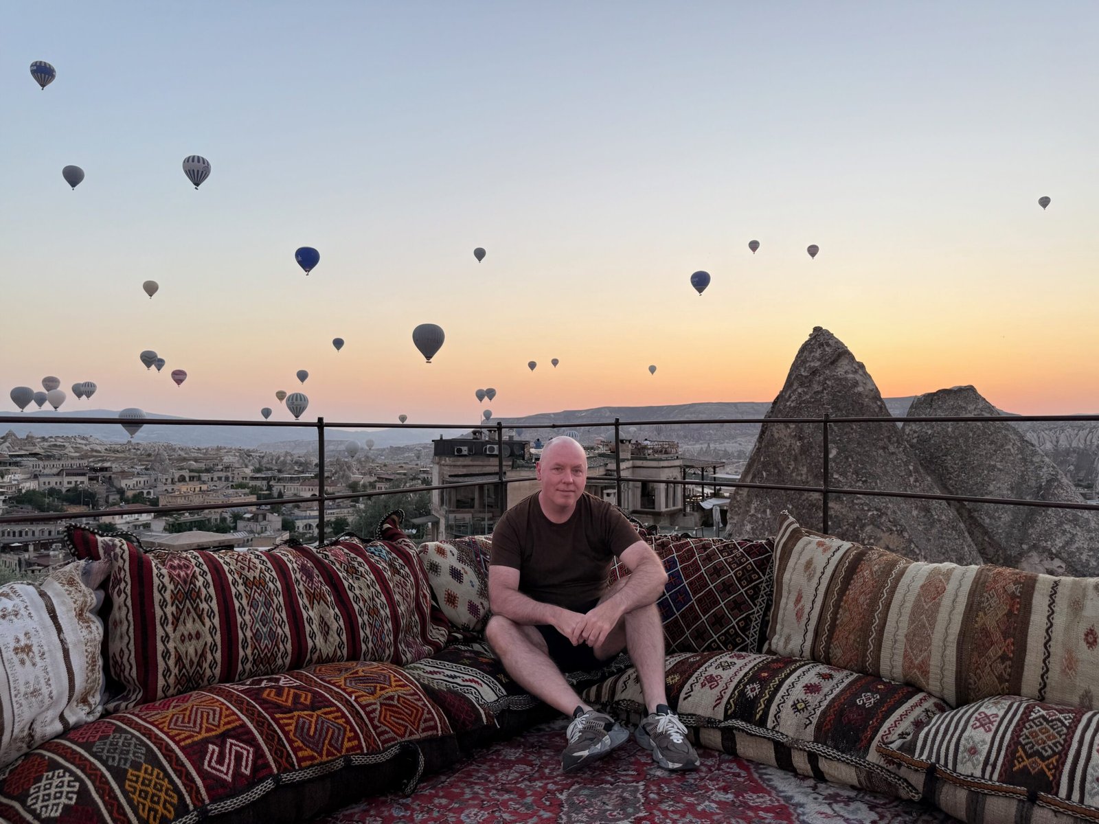 Author Don Trynor sitting on a rooftop terrace in Göreme, Türkiye, with dozens of Cappadocia hot air balloons floating at dawn above the valleys and fairy chimneys.