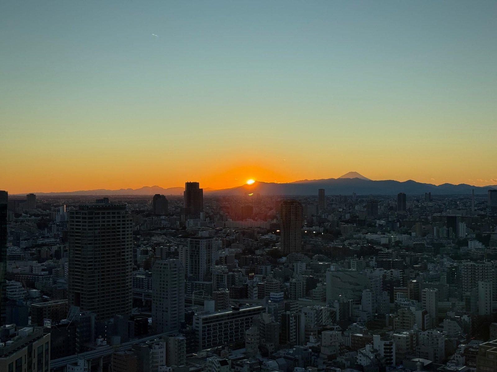 Tokyo Tower sunset showing the sun touching the distant mountains, with Mount Fuji on the horizon and the city skyline below.