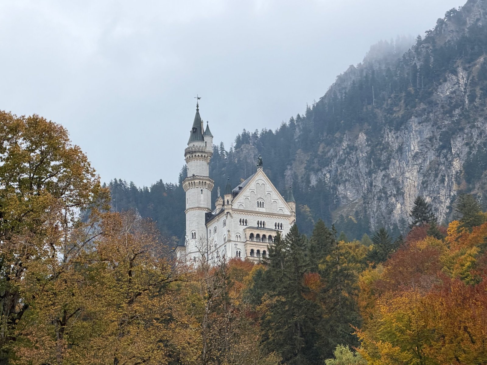 Neuschwanstein Castle viewed from the hiking trail near Hohenschwangau, surrounded by autumn trees with limestone cliffs rising behind.