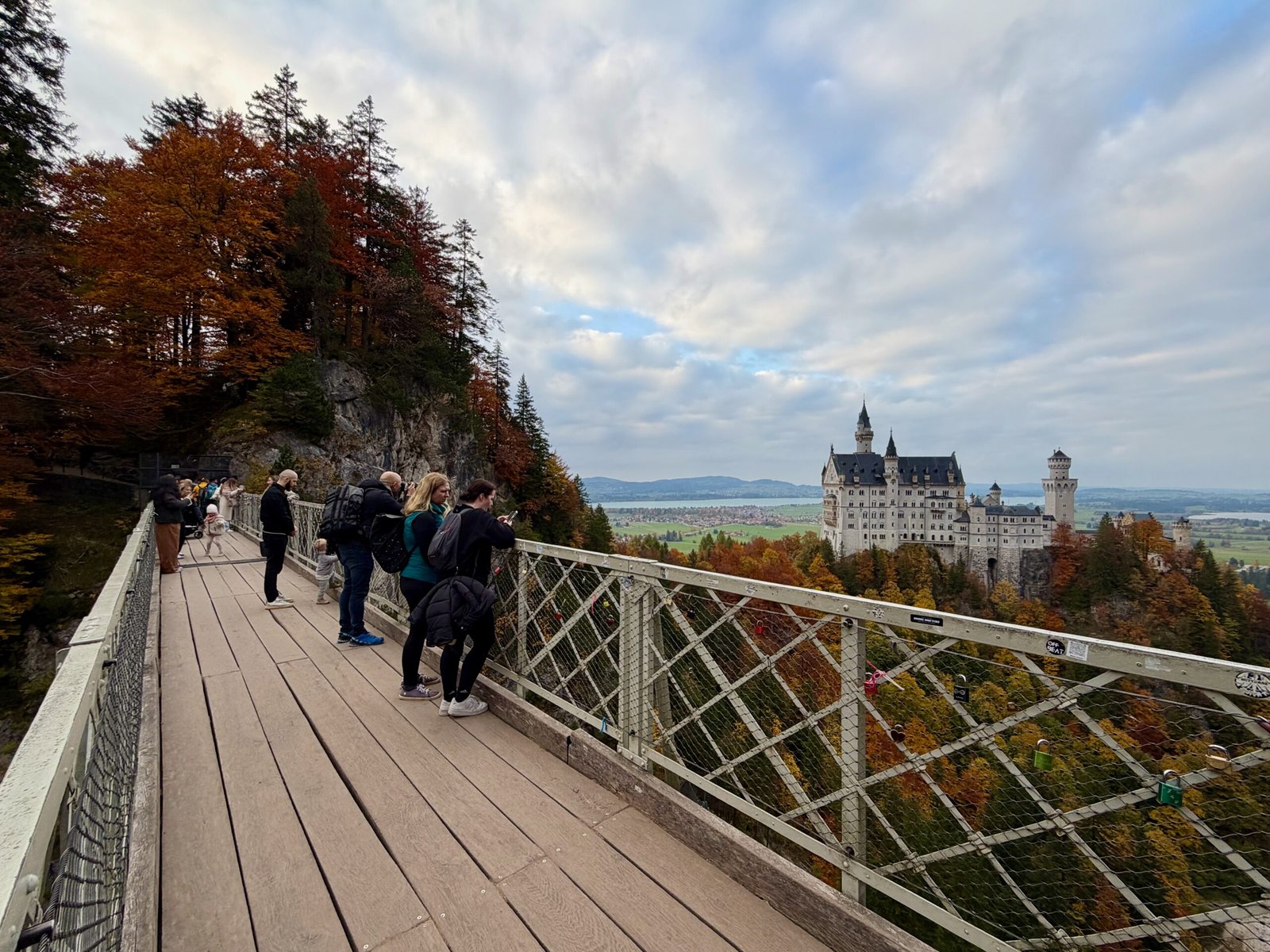 Visitors on Marienbrücke bridge over the Pöllat Gorge admiring Neuschwanstein Castle in the Bavarian Alps, surrounded by autumn forest.